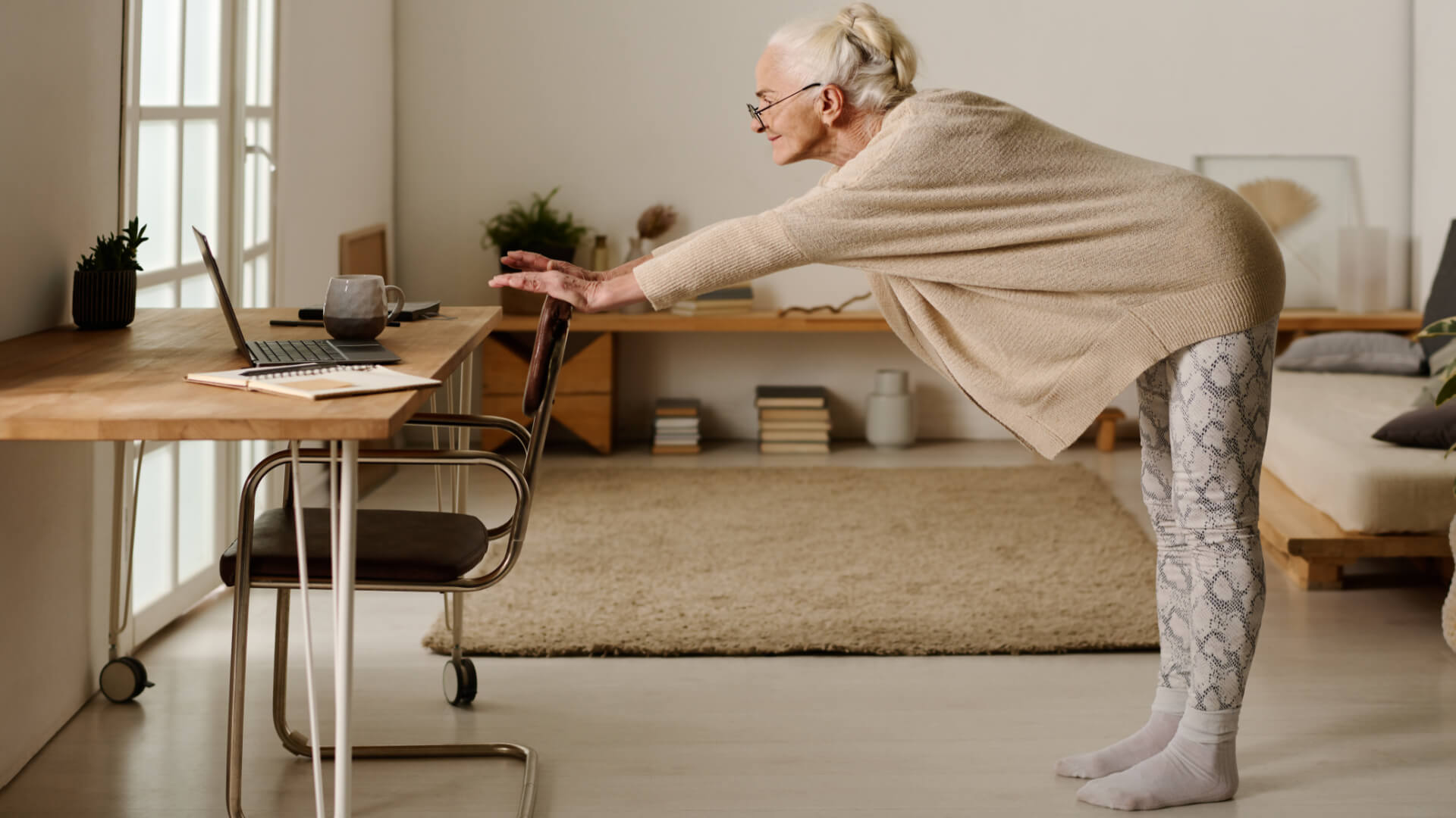 elderly woman wearing leggings and a beige cardigan with grey hair and glasses is holding onto a the back of a chair exercising while looking at her laptop. 