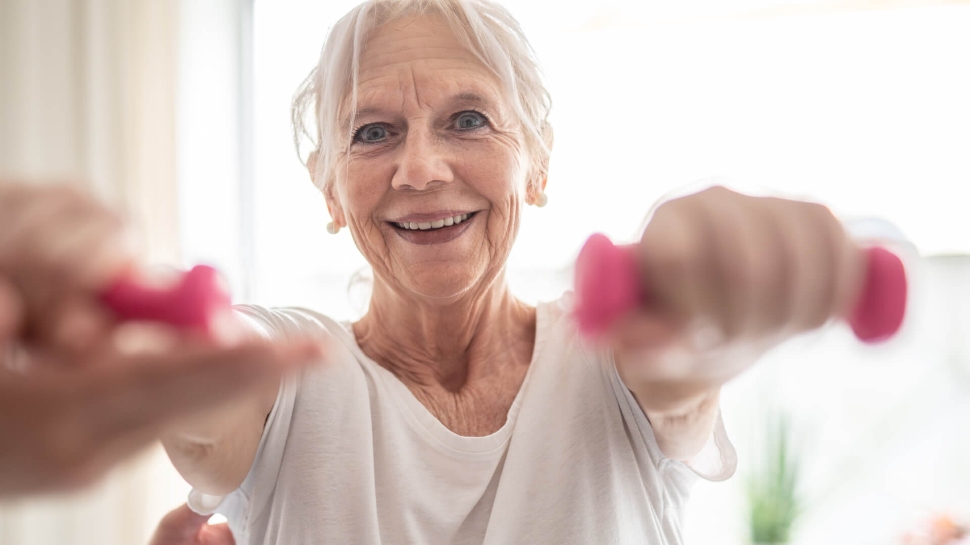 Elderly silver haired woman wearing a white t-shirt engaging in rehabilitative exercises with pink small dambbells in her hands