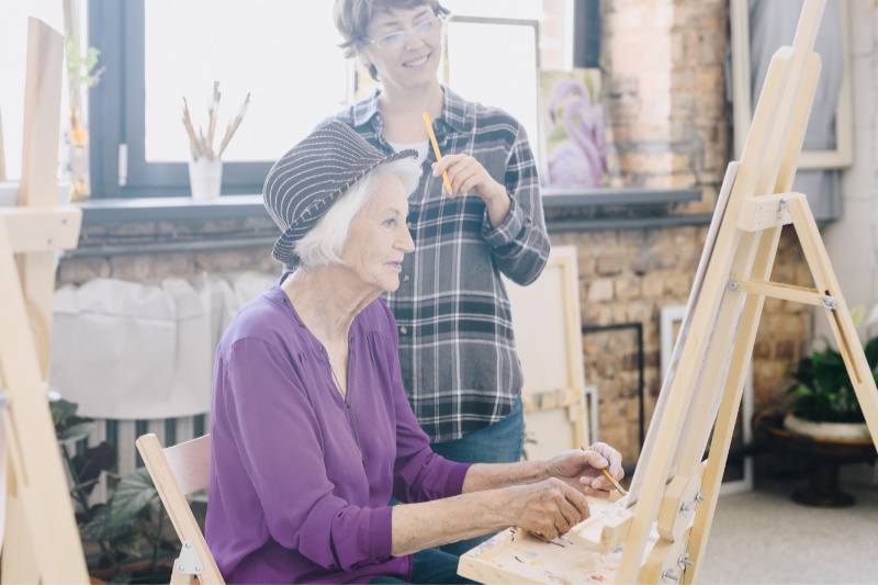 Side view portrait of elegant senior woman painting sitting at easel in art studio studying art with smiling female teacher giving comments, scene in spacious sunlit loft space.