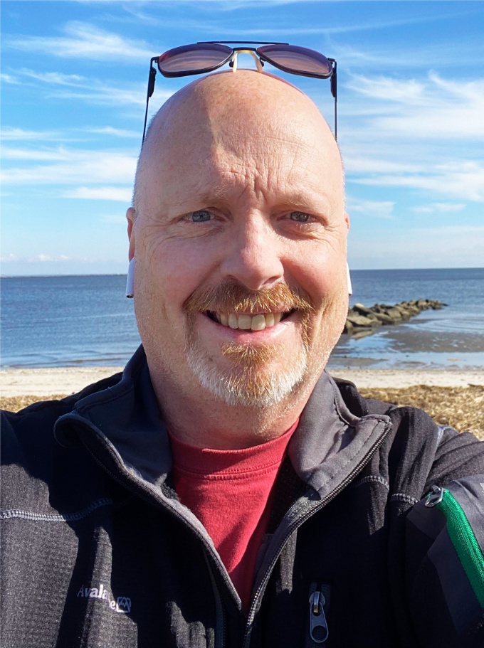 bald middle aged man with short white beard, smiling at the beach with ocean behind him, wearing a dark grey jacket.